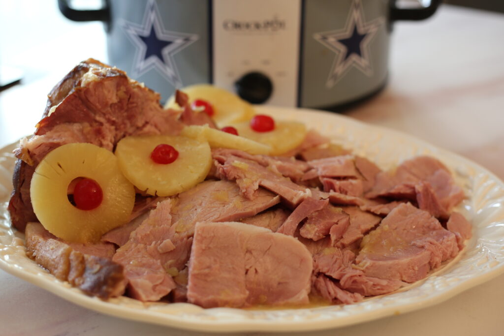 sliced ham with pineapple slices and red cherries on a platter next to a crockpot