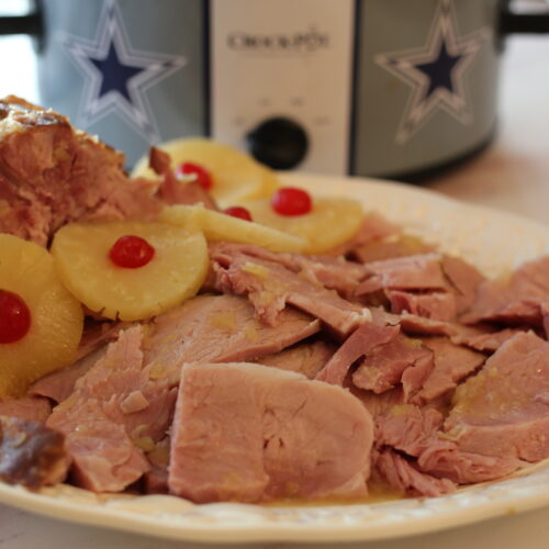 sliced ham with pineapple slices and red cherries on a platter next to a crockpot