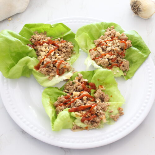 Three lettuce wraps filled with ground chicken on a white plate. Topped with red hot sauce.