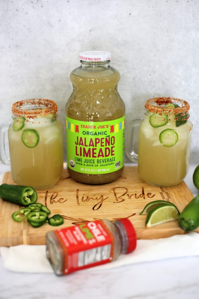 Jalapeño limeade jar with two glasses of margaritas next to it on a brown cutting board.